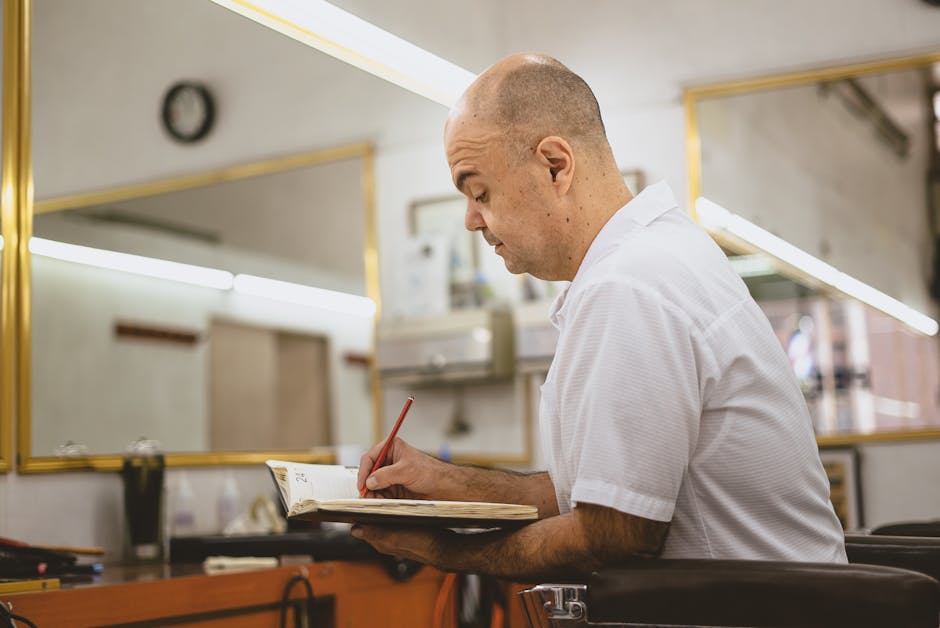 Bald man writing in a notebook inside a modern barber shop with mirrors.